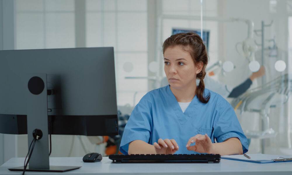 Stomatological assistant typing on computer keyboard sitting at oral clinic desk. Nurse using technology while professional orthodontist consulting patient with equipment in background